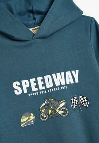 Sudadera color verde azulado con un gráfico frontal que presenta "SPEEDWAY", una motocicleta, un casco y banderas a cuadros. Material de algodón suave, diseño casual.