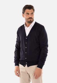 Cardigan in lana blu navy con scollo a V, chiusura a bottoni, polsini e vita a coste. Presenta una superficie liscia e una leggera lucentezza. Indossato sopra una camicia bianca.