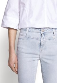 Jeans de mezclilla azul claro con un ajuste slim, diseño de cinco bolsillos y cierre de botón plateado, combinados con una camisa blanca de manga corta.