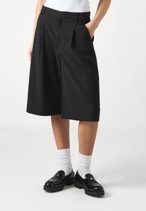 Person iført sorte vidde-benskorts til knæet med plisseringer, hvide sokker, sorte loafers og en hvid top, stående med den ene hånd i lommen.