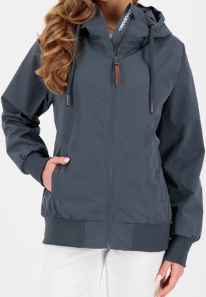 Vrouw die een donkergrijze zip-up capuchontrui draagt met trekkoorden, geribbelde boorden en zoom, handen in de met knopen afgesloten voorkantzakken, gecombineerd met een witte broek.