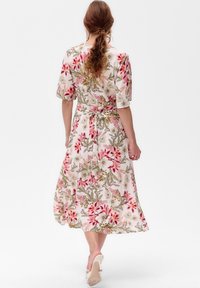 Frau mit langen lockigen Haaren, die ein florales Midikleid mit rosa und grünen Mustern trägt, geht in weißen High Heels davon.