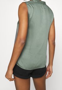 Ärmelloses grünes Shirt aus leichtem Stoff, mit einem Knopfverschluss am Rücken, leicht abgerundetem Saum und lässiger Passform.