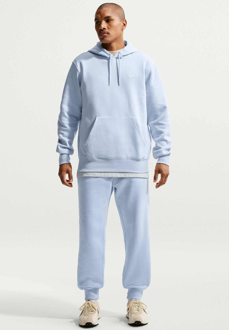 Uomo in piedi che indossa una felpa con cappuccio azzurro chiaro e pantaloni della tuta abbinati con sneakers bianche, su uno sfondo chiaro e semplice.