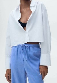 Bílá zkrácená blůza s knoflíkovou přední stranou, dlouhými rukávy a volným střihem, v kombinaci s světle modrými kalhotami na šňůrku. Černá podprsenka je viditelná pod blůzou.