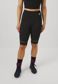 Sorte sykkelshorts med mesh-detaljer nær sømmen, høy midje-design, sammen med sorte atletiske sko med røde detaljer.