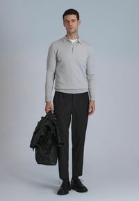 Helles graues, geripptes Langarmpolo mit Kragen, kombiniert mit schwarzen, maßgeschneiderten Hosen und schwarzen Schnürschuhen, während er eine schwarze Pufferjacke hält.