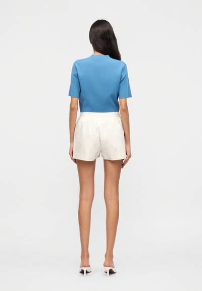 Femme debout de dos, portant un haut bleu à manches courtes, un short blanc à motifs floraux et des mules blanches à talons sur un fond blanc uni.