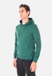 Giovane uomo con felpa verde a cappuccio con tasca frontale e pantaloni cargo neri, in piedi e che guarda leggermente a sinistra su uno sfondo bianco.