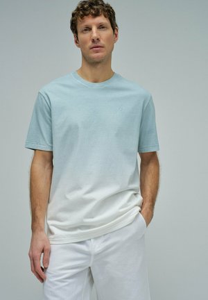 Hombre con camiseta de manga corta ombré en azul claro y blanco y pantalones blancos, de pie frente a un fondo gris liso, con las manos en los bolsillos.