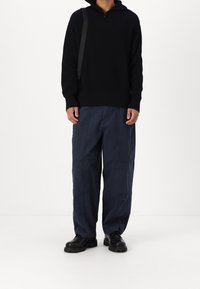 Personne portant un pull en maille noire, un pantalon ample bleu foncé, des chaussures en cuir noires et un sac à bandoulière noir sur un fond blanc uni.