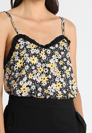 Bluzka bez rękawów w kwiaty żółte i białe na czarnym tle, z cienkimi ramiączkami i falbankowanym dekoltem, noszona z czarnymi spodniami.