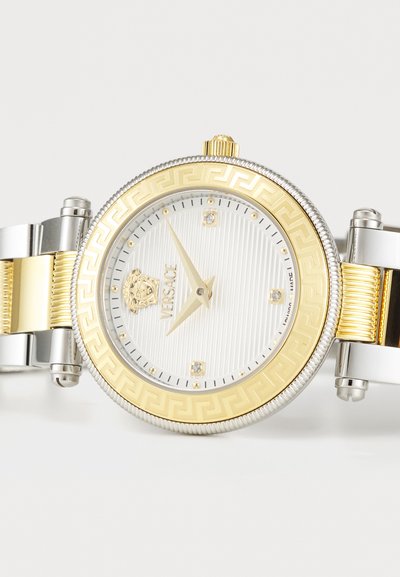 Gold and silver metal watch with a round face, textured white dial, diamond accents, and a Greek key pattern on the bezel.