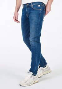 Jeans in denim blu con una vestibilità slim, dotati di cinque tasche, chiusura con bottone e una piccola etichetta logo sulla cintura. Abbinati a scarpe da ginnastica grigie e bianche.