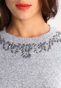Ljusgrå tröja med en texturerad yta och ett dekorativt blomstermönster med pärlor längs halsringningen i olika nyanser av grått.