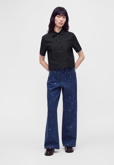 Femme aux longs cheveux foncés portant une chemise noire à manches courtes boutonnée et un jean bleu à jambes larges avec de petits motifs blancs, debout les mains dans le dos.