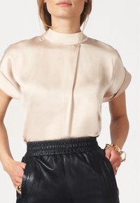 Seidenähnliches, hellbeiges Oberteil mit hohem Kragen und kurzen Ärmeln, kombiniert mit schwarzen Lederhosen mit elastischem Bund.