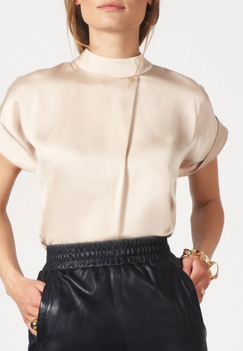 Seidenähnliches, hellbeiges Oberteil mit hohem Kragen und kurzen Ärmeln, kombiniert mit schwarzen Lederhosen mit elastischem Bund.