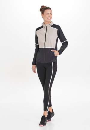 Kevyt takki beigen ja mustan värisenä, jossa on vetoketju edessä, huppu ja heijastavat yksityiskohdat; yhdistetty mustiin leggingsihin, joissa on valkoiset raidat.