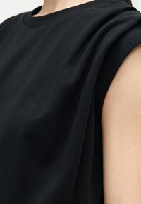 Close-up of a person wearing a sleeveless black top, showing folded fabric details on the shoulder and upper arm exposed.