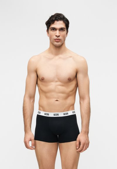 Άνδρας χωρίς πουκάμισο με κοντά σκούρα μαλλιά, φορώντας μαύρα εσώρουχα μποξερ με λευκή μέση που φέρει την επιγραφή "GCDS", στέκεται μπροστά σε ένα απλό φόντο.