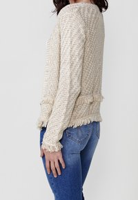 Beżowy dziany kardigan o fakturowanym splocie, z frędzlami na dole i przy mankietach, otwarty z przodu, noszony na niebieskich dżinsach.