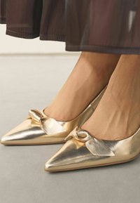 Zelta, metāla, ar smailu purngalu kurpes ar satīna loku priekšpusē. Gluda faktūra, kas atspoguļo gaismu, uz gaiši pelēka virsma.