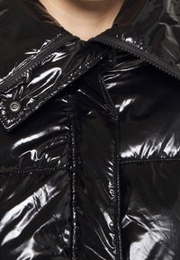 Sort pufferjakke med en blank finish, oversized krave og synlige syninger. Lukkes med en lynlås og knapper foran.