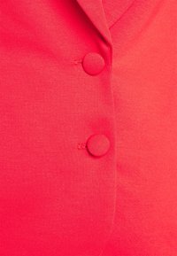 Vêtement boutonné corail vif avec un tissu texturé, un col en V et deux boutons recouverts du même tissu à l’avant.