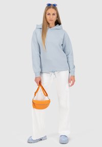 Felpa leggera blu con coulisse, pantaloni bianchi a gamba ampia e borsa arancione; il modello indossa occhiali da sole blu e calzature chiare.