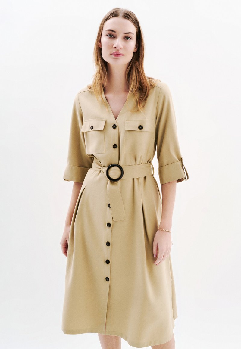 Vestito khaki con bottoni frontali, maniche corte, due tasche sul petto, una cintura in vita con una fibbia rotonda e una silhouette ampia e fluida.