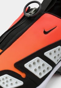 Nærbilde av oransje og svarte Nike-sko med glidelås og sirkulært trekk på den øvre delen av skoen.