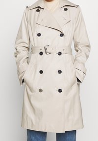 Beżowy trencz z dwurzędowym zapięciem, czarnymi guzikami, paskiem w talii i bocznymi kieszeniami. Posiada pagony na ramionach oraz gładką fakturę.