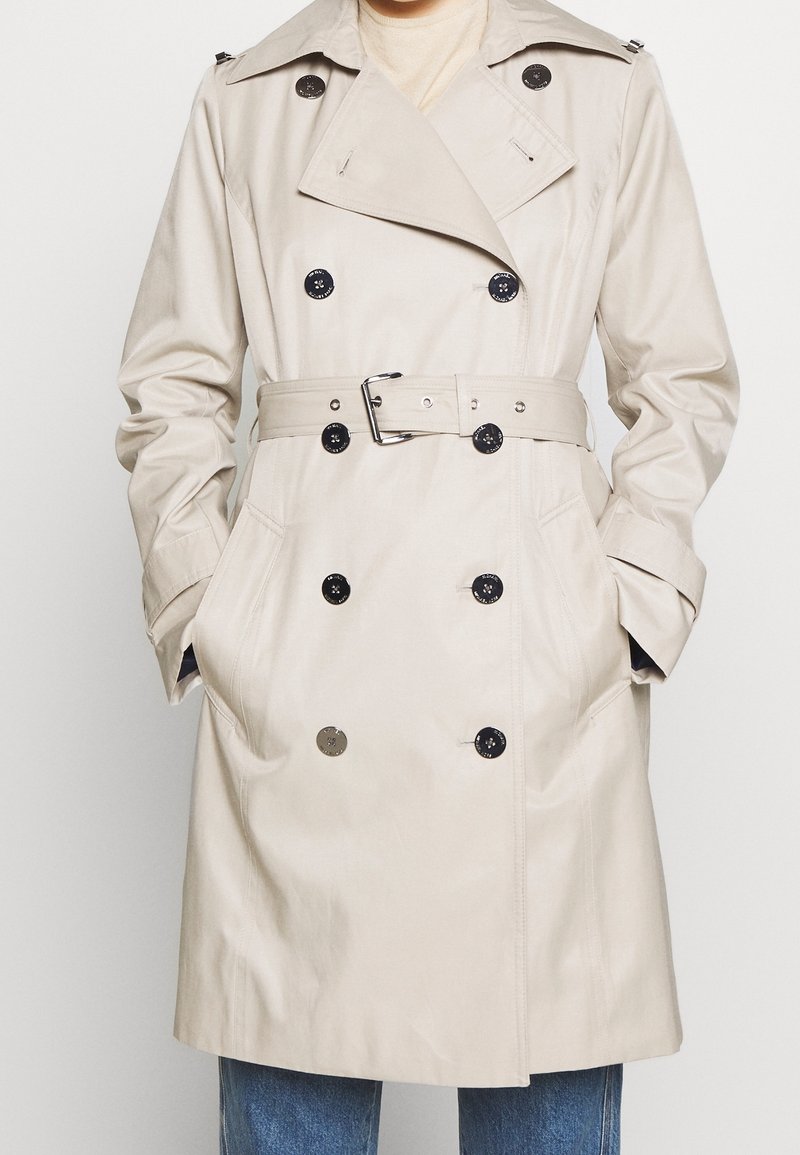 Beżowy trencz z dwurzędowym zapięciem, czarnymi guzikami, paskiem w talii i bocznymi kieszeniami. Posiada pagony na ramionach oraz gładką fakturę.