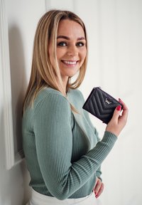 Black quilted wallet with chevron pattern, silver hardware logo, held in hand. Light green ribbed long-sleeve top against a white wall.