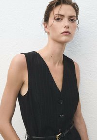 Black pinstripe sleeveless dress with a v-neck, featuring a button-down front and a slim belt with a gold buckle. Smooth fabric texture.