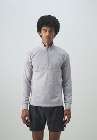 Ljust grå atletisk pullover med hög krage, kvartdragkedja och kontrasterande texturerad tyg över bröstet. Kombinerad med mörka shorts.