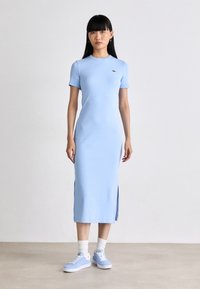 Robe bleu clair mi-longue avec des manches courtes, des fentes latérales et un petit logo. Associée à des baskets bleu clair et des chaussettes blanches. Texture lisse.