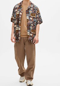 Chemise à manches courtes à motifs abstraits en brun, bleu et beige, associée à un pantalon marron et des baskets blanches.