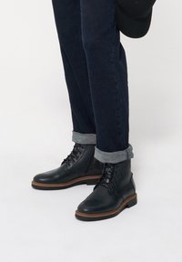 Czarne skórzane botki za kostkę z okrągłym noskiem i sznurowanym zapięciem, z brązową gumową podeszwą. W połączeniu z ciemnymi dżinsami i podwiniętymi mankietami.