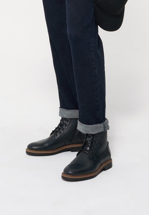 Czarne skórzane botki za kostkę z okrągłym noskiem i sznurowanym zapięciem, z brązową gumową podeszwą. W połączeniu z ciemnymi dżinsami i podwiniętymi mankietami.