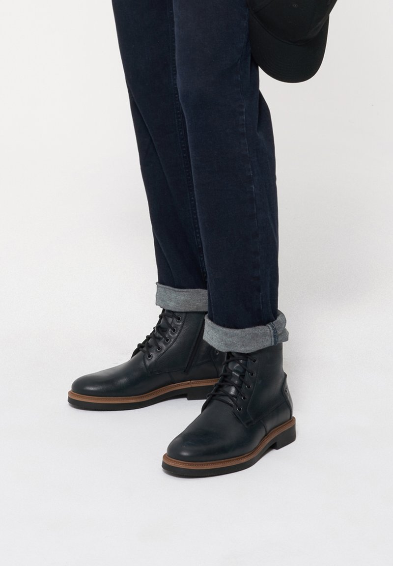 Czarne skórzane botki za kostkę z okrągłym noskiem i sznurowanym zapięciem, z brązową gumową podeszwą. W połączeniu z ciemnymi dżinsami i podwiniętymi mankietami.