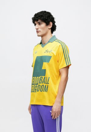Young man with curly hair wearing a yellow football jersey with green text and purple pants, standing against a plain white background.