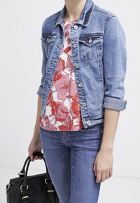 Giacca di jeans azzurro chiaro con un design aderente, tasche frontali e maniche arrotolate. Indossata sopra un top con fantasia floreale e abbinata a jeans.