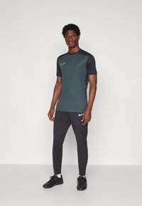 Športový outfit Nike, ktorý obsahuje tmavozelené tričko s krátkym rukávom, čiernymi doplnkami a žltým logom, kombinované s čiernymi nohavicami a čiernymi topánkami.