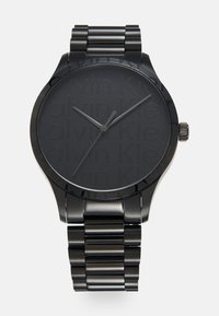 Reloj de metal negro con acabado mate, que presenta una esfera redonda y un dial texturizado grabado con el nombre de la marca. Pulsera de eslabones metálicos.