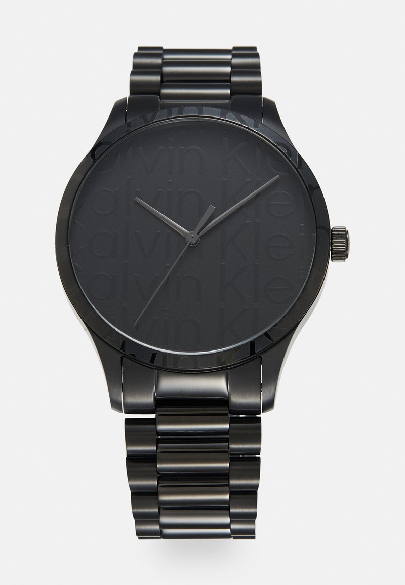 Montre noire en métal avec une finition mate, un cadran rond et un dial texturé embossé du nom de la marque. Bracelet en maillons métalliques.