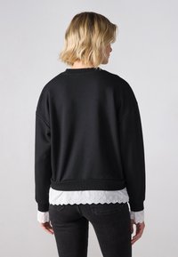 Felpa nera con colletto e polsini a coste, caratterizzata da un dettaglio a strati di pizzo bianco sul fondo. Il materiale appare morbido e con una texture interessante.