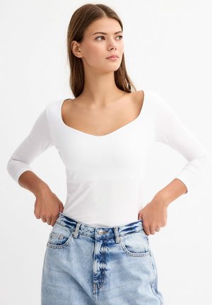 Top blanco de manga larga con un escote amplio, combinado con pantalones cortos de mezclilla azul claro de cintura alta, que muestra un ajuste relajado y una textura suave.