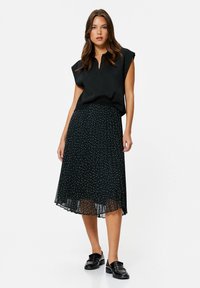 Sort ærmeløs top parret med en sort plisseret nederdel med hvide polka dots. Modellen bærer sorte loafers, der viser et lagdelt design.